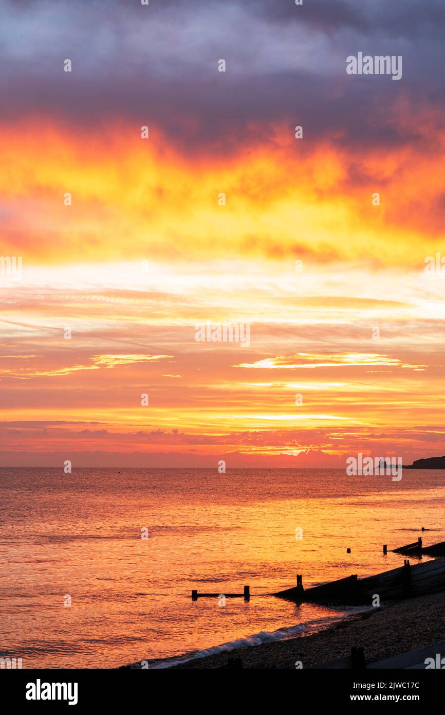 The dawn sky over the sea on the Kent coast at Reculver with the twin towers of the 12th century church silhouetted in the distance against an orange sky. In the foreground the silhouetted breakwaters, groynes, and beach at Henre Bay. Thick cloud overhead underlit by orange light. Stock Photo
