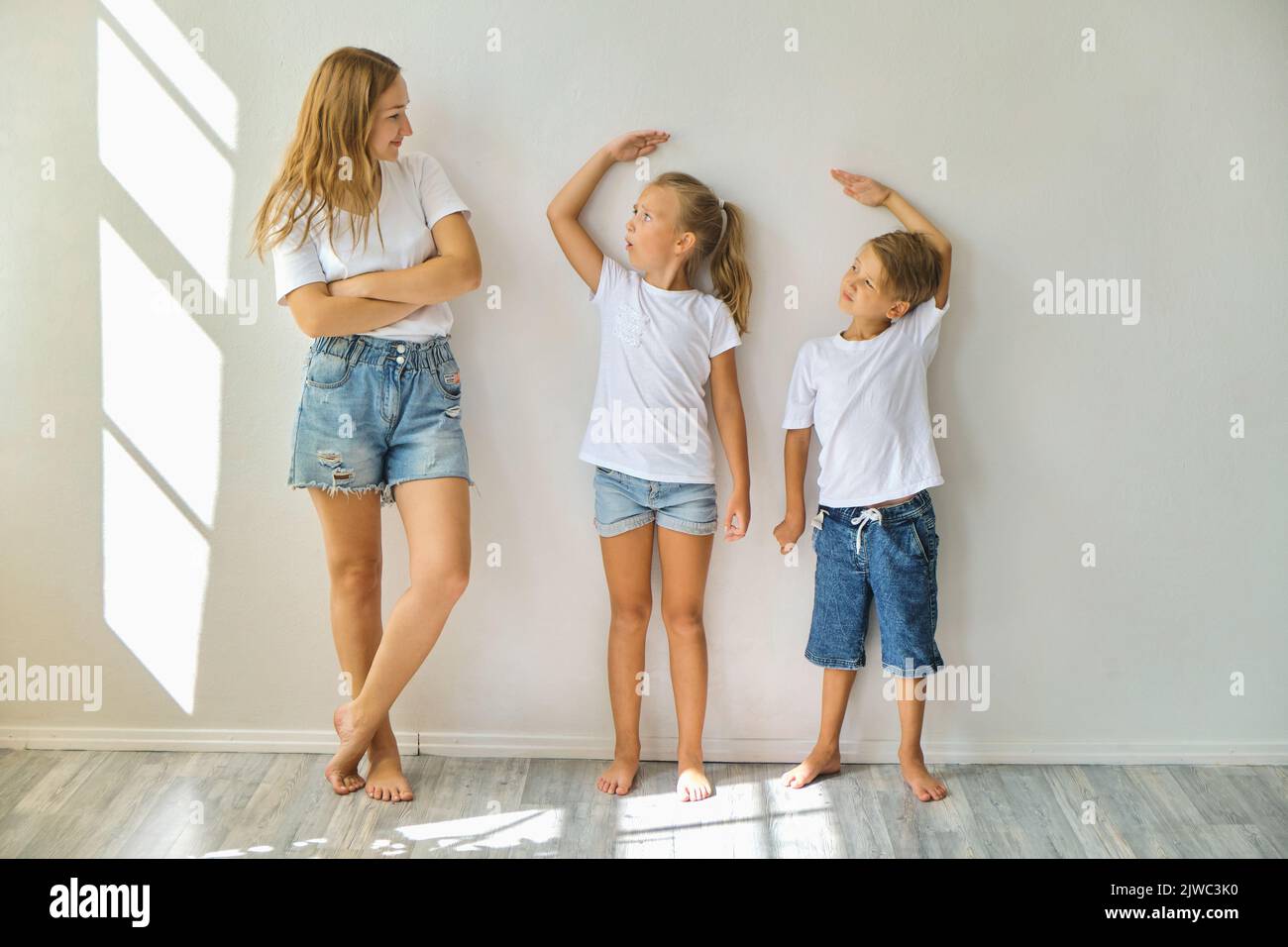 mother measures growth of kids to a daughter and son near light grey wall Stock Photo