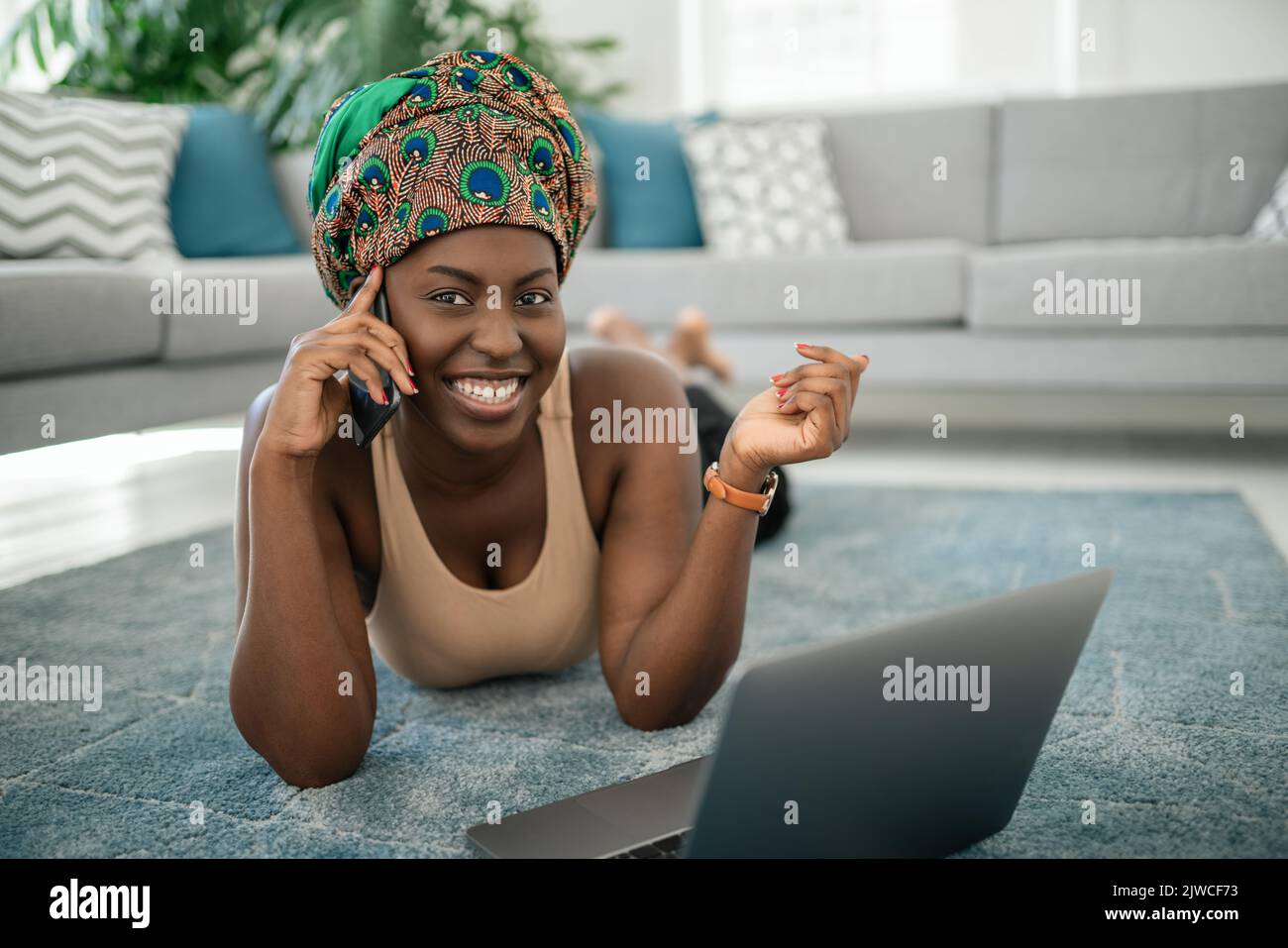 Beautiful African woman at home, smiling and laughing looking into camera, using mobile smart phone, wearing traditional headscarf, surprised and happ Stock Photo