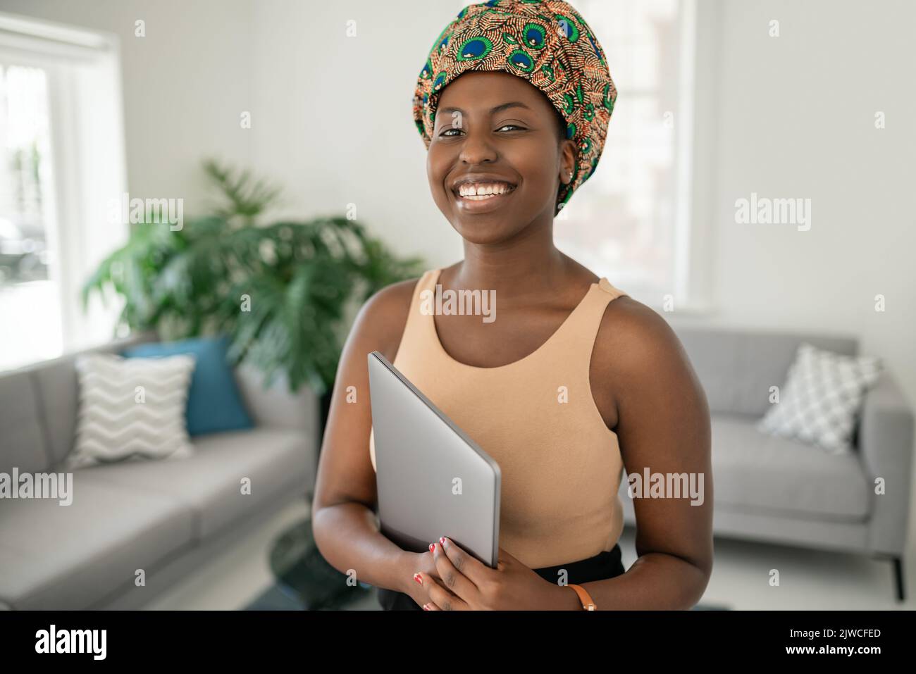 Portrait of beautiful smiling African woman standing in modern home with laptop in hand, wearing a traditional headscarf Stock Photo