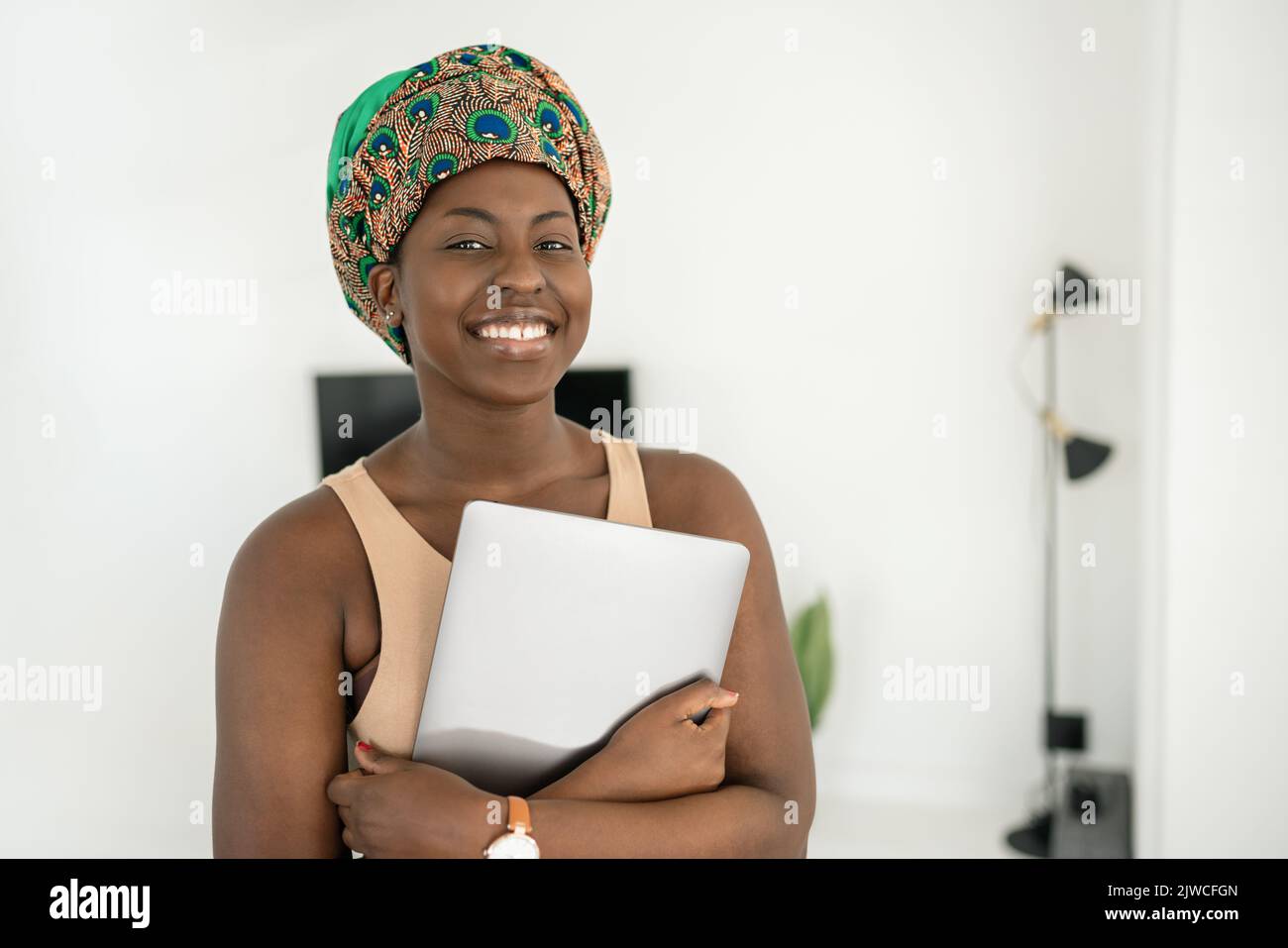 Portrait of beautiful smiling African woman standing in modern home with laptop in hand, wearing a traditional headscarf Stock Photo