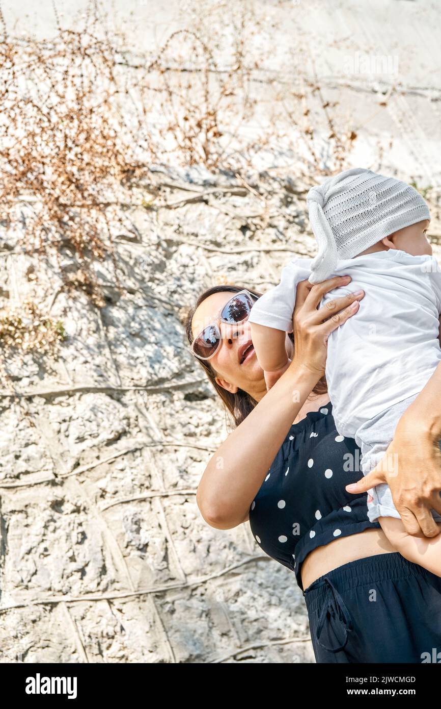Mother in sunglasses holds cute toddler in arms against the wall. Young brunette woman and little boy enjoy summer vacation low angle close view Stock Photo
