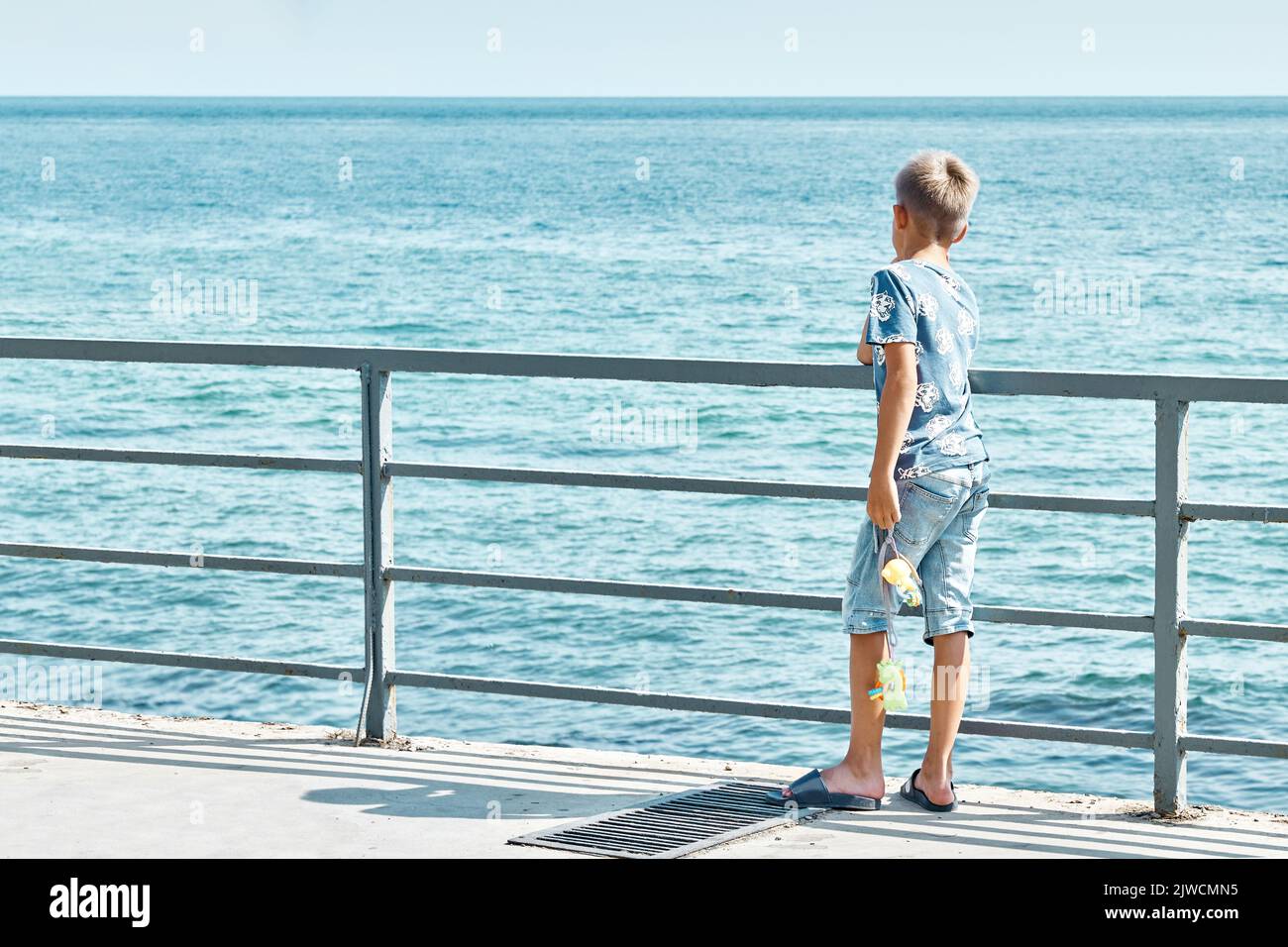 Junior schoolboy stands on pier looking at seascape at bright sunlight. Schoolchild enjoys holidays travelling at seaside. Light waves hit pier Stock Photo