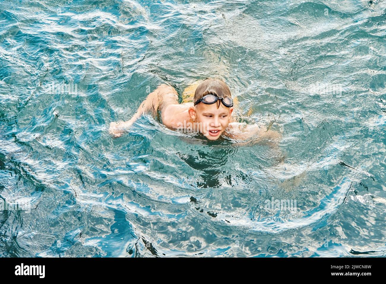 Junior schoolboy in swimming goggles enjoys holidays. Schoolkid swims in clear blue sea with smiling and excited expression on face closeup Stock Photo