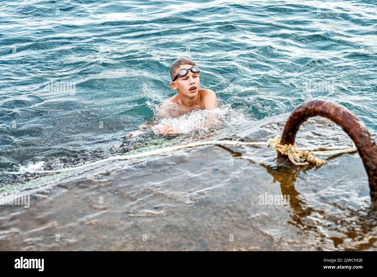 Schoolboy catches and holds rope swimming in blue water trying to climb up stone pier. Guy wearing swimming goggles spend vacation at seaside Stock Photo