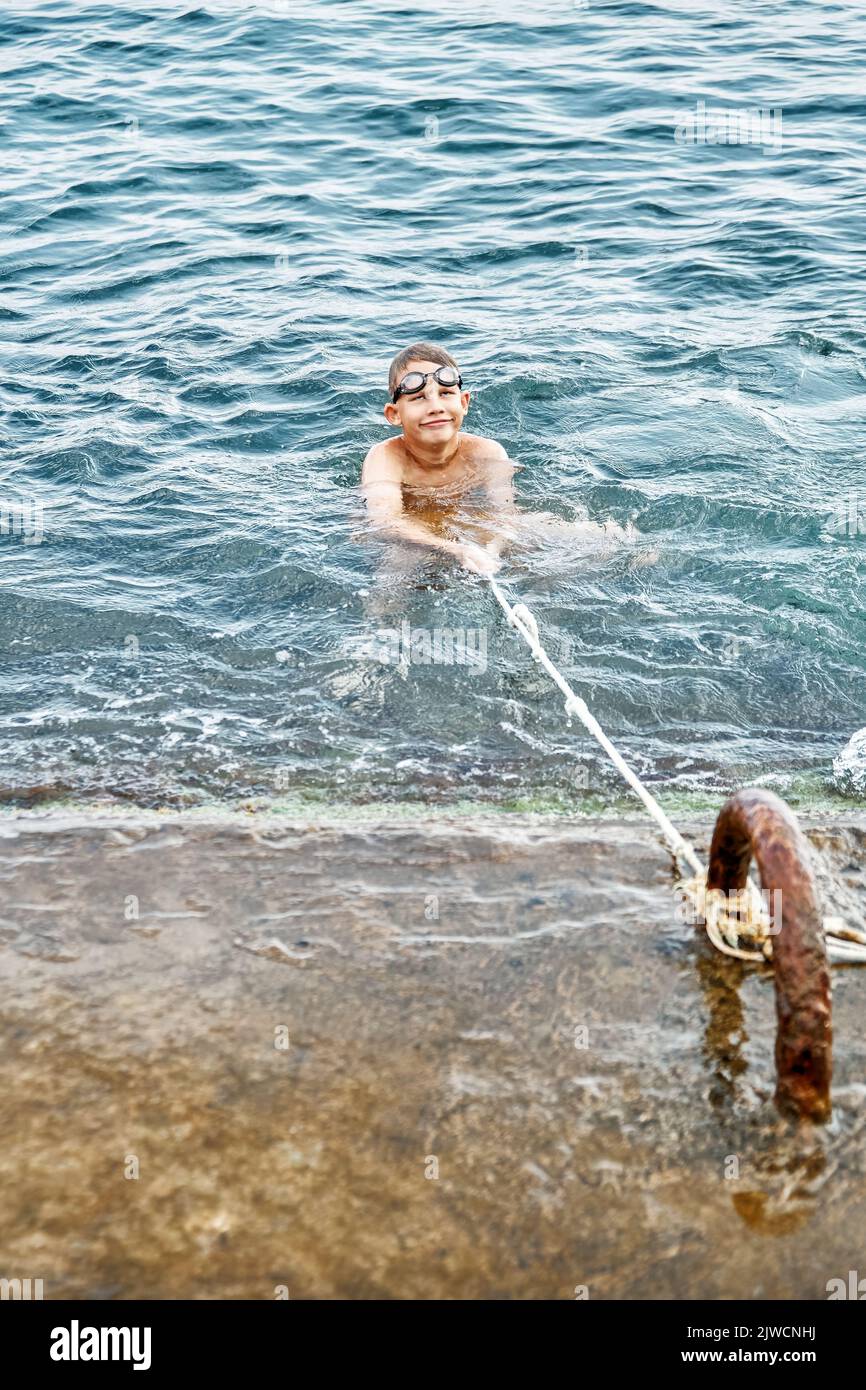 Schoolboy catches and holds rope swimming in blue water trying to climb up stone pier. Guy wearing swimming goggles spend vacation at seaside Stock Photo