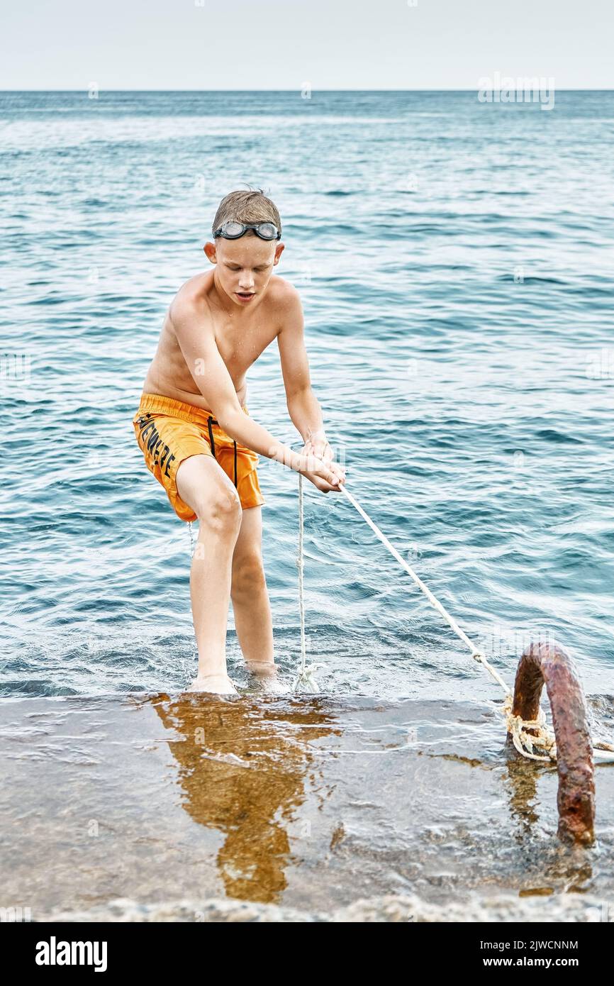Junior schoolboy wearing yellow swimming trunks and goggles climbs up pier holding rope against blue seascape. Boy enjoys holidays at sea closeup Stock Photo