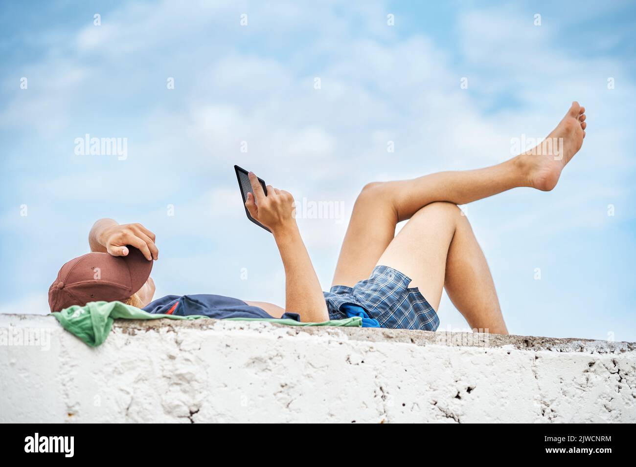 Young guy reads digital book on tablet lying on stone pier with bare legs against blue sky. Young lad enjoys reading uniting with nature at seaside Stock Photo