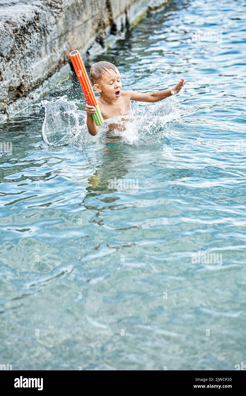 Moment of small boy with water-jet gun diving into water while jumping in sea from pier. Small kid jumps and splashes water at summertime Stock Photo