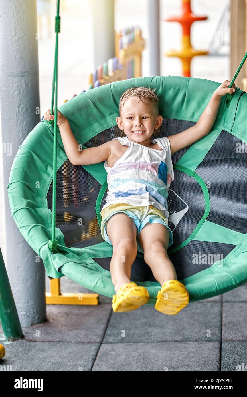 Small blond boy sits in green swing armchair at coastal resort. Small child swings on hotel children playground enjoying happy holidays at seaside Stock Photo