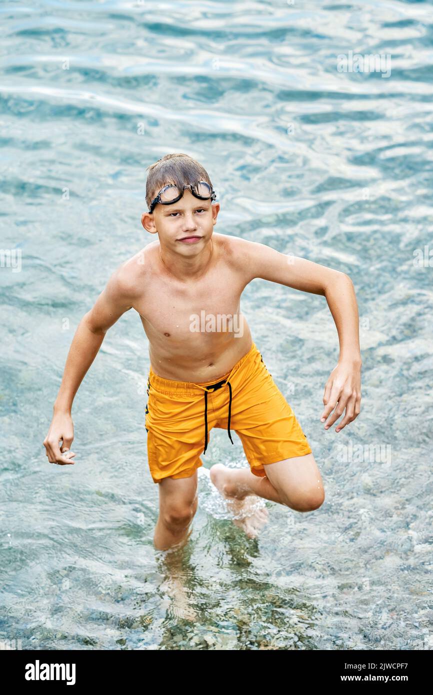 Happy schoolboy wearing yellow trunks and swimming goggles walks wet from water to beach grimacing. Boy enjoys summer vacation closeup Stock Photo