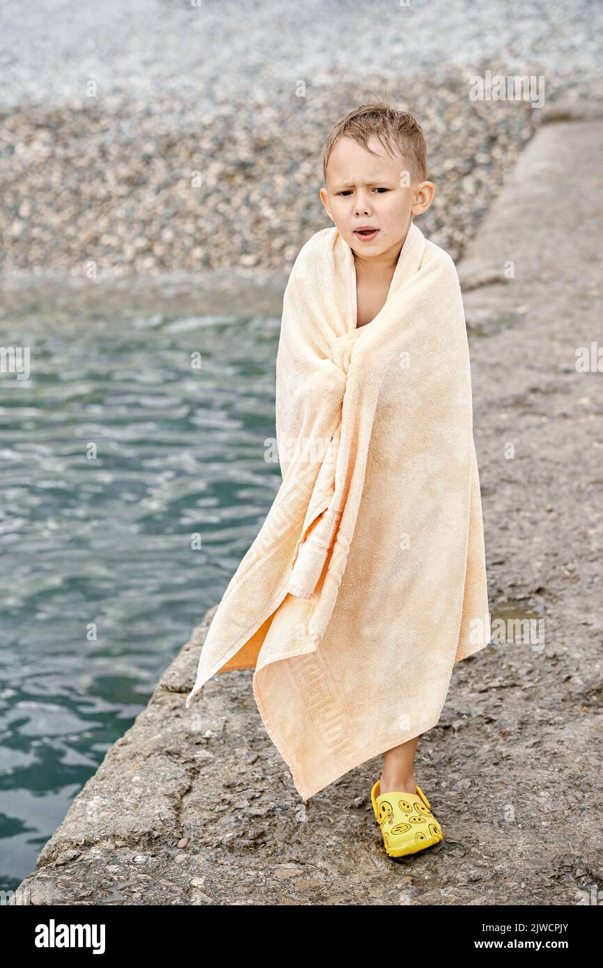 Small boy wrapped in beige terry towel shivers from cold after swimming in sea. Preschooler boy tries to warm up with huge towel on stone pier closeup Stock Photo