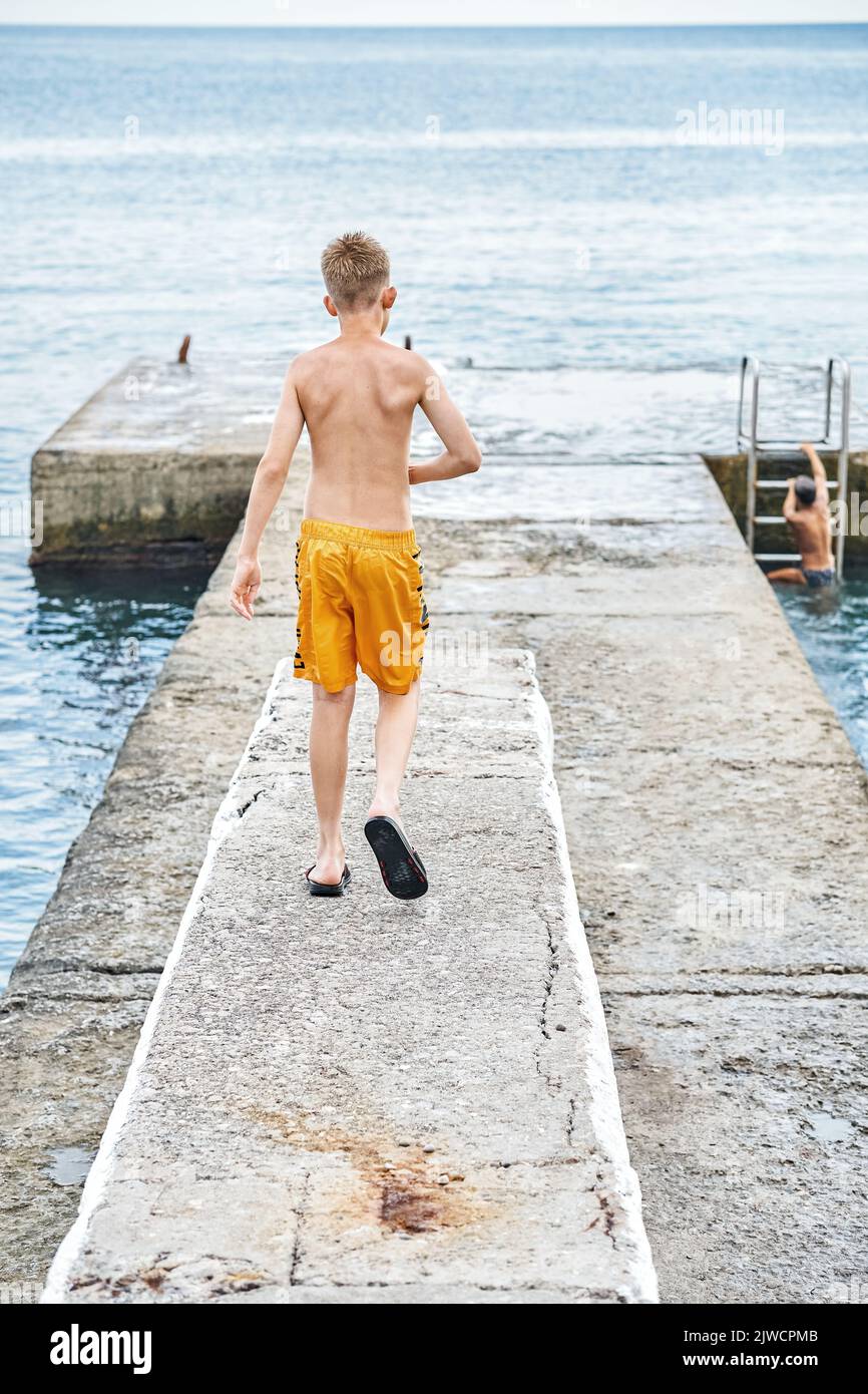 Moment of schoolboy walks on stone pier watching seascape. Young guy wearing yellow trunks wants to jump into sea spending summer holidays at seaside Stock Photo