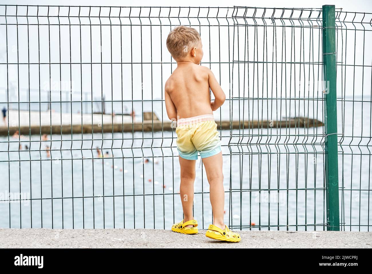 Small boy in shorts stands on parapet looking at seascape behind lacy fence in morning. Young boy enjoys summer holiday at seaside closeup Stock Photo
