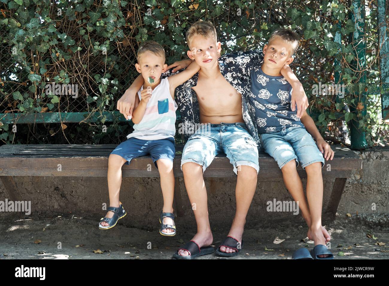 Friendly siblings sit on stone bench resting in tree shadow on summer holidays. Three blond brothers in shorts enjoy spending time together Stock Photo