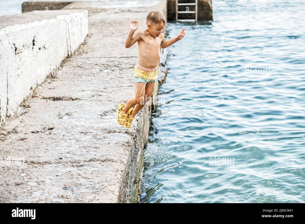 Small boy jumps flying from pier in funny posture into sea water. Preschooler child enjoys spending holidays with family at seaside Stock Photo