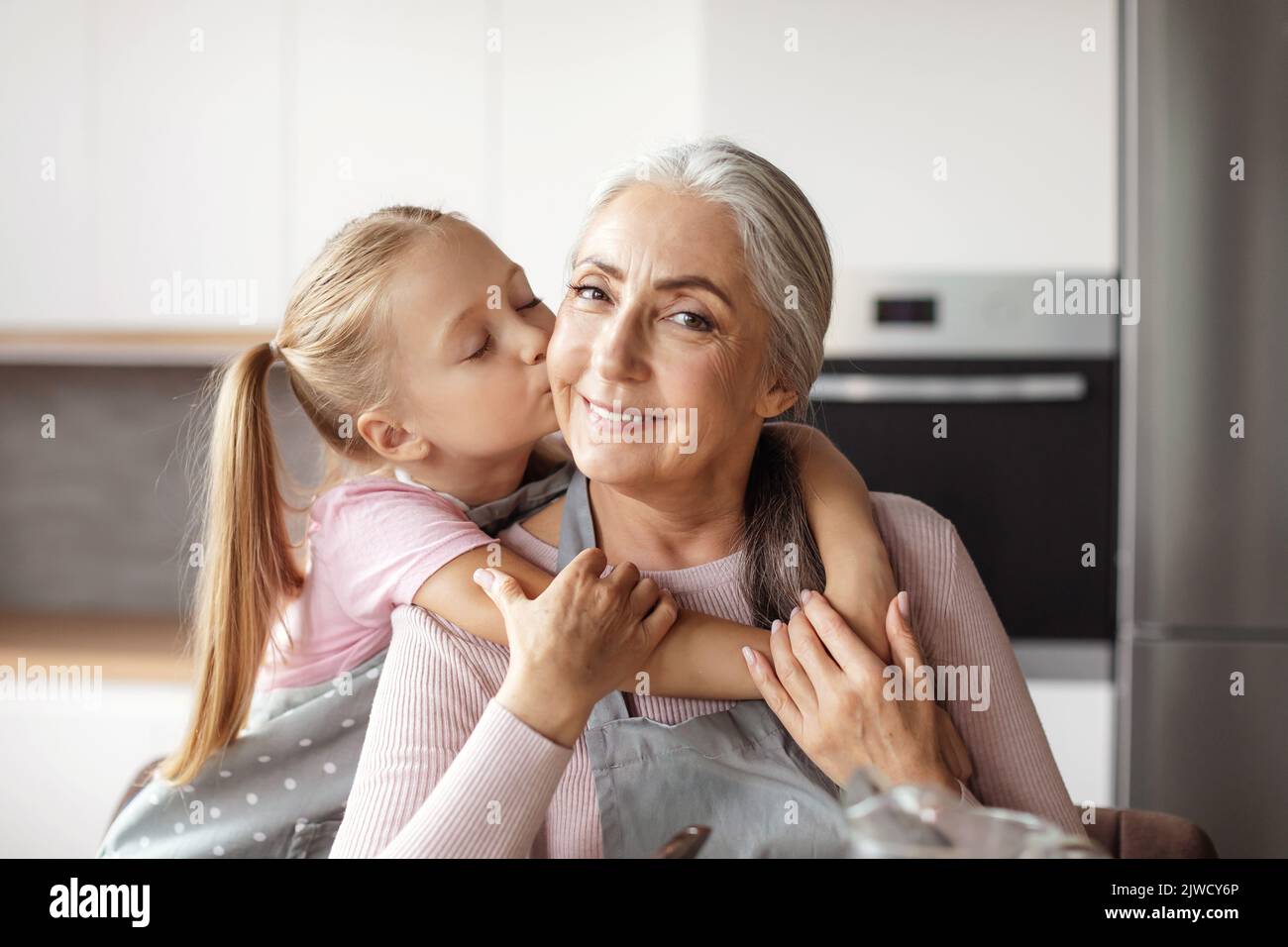 Portrait of smiling european little granddaughter kisses cheek of elderly grandmother in apron Stock Photo