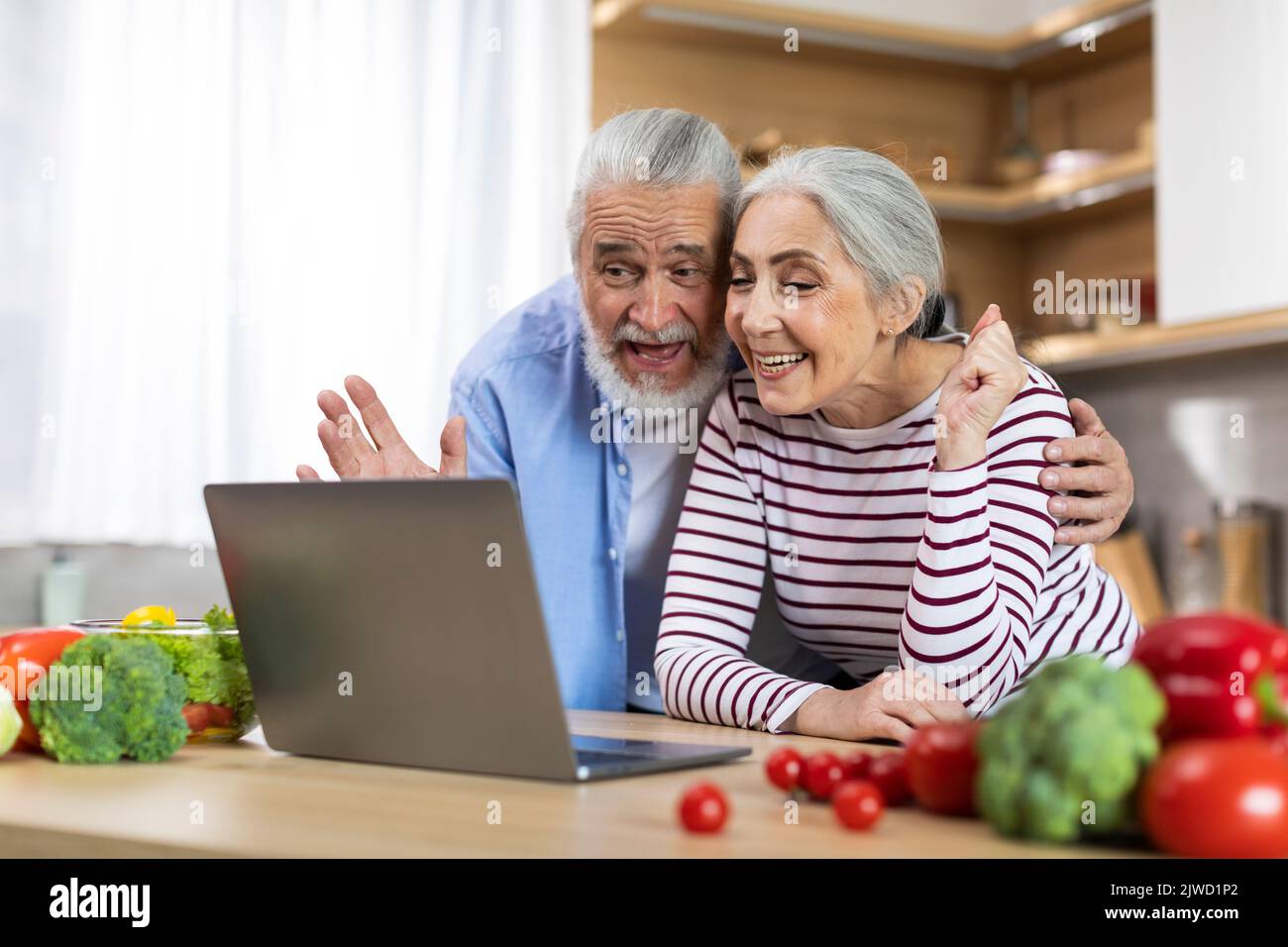 Happy Senior Couple Making Video Call With Laptop While Relaxing In Kitchen Stock Photo
