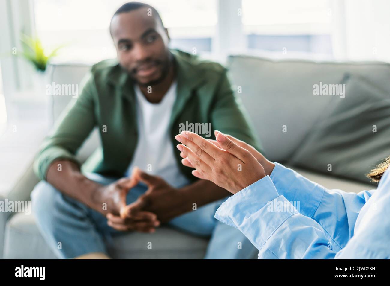 Pensive black man sitting on sofa at psychotherapist cabinet and listening to specialist, focus on psychologist hands Stock Photo