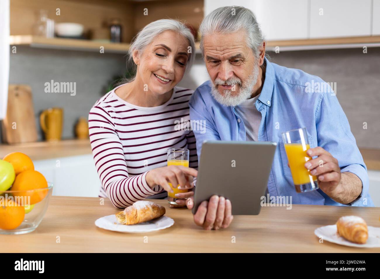 Cheerful Elderly Couple Having Breakfast Together And Using Digital Tablet In Kitchen Stock Photo