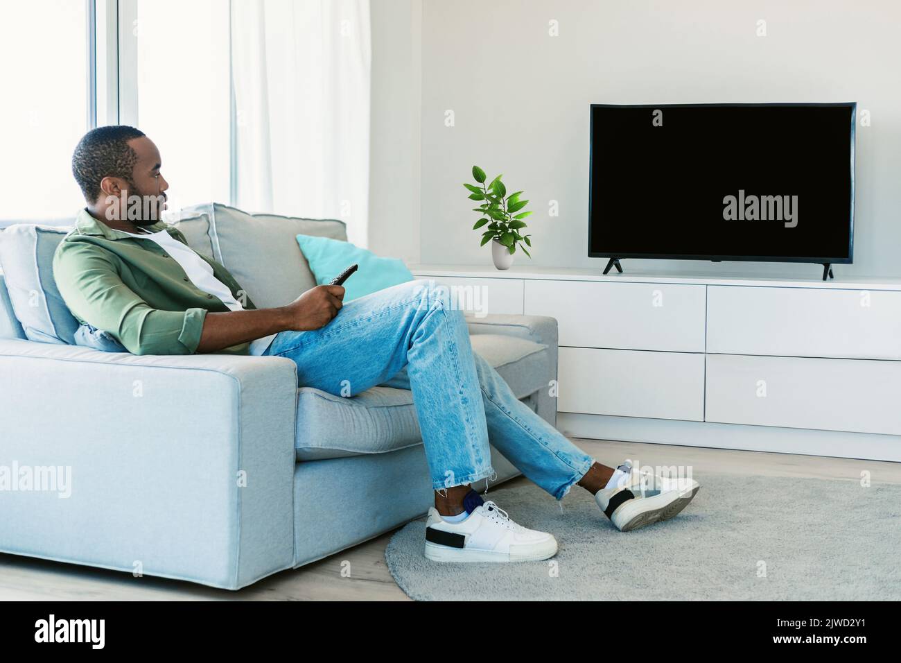 Rest and relax. Black man sitting on the sofa in living room, watching television show, looking at blank plasma tv Stock Photo