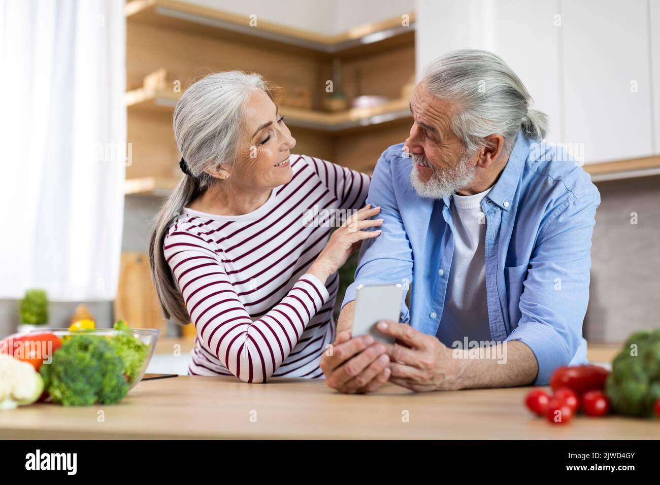 Modern Technologies. Happy Senior Spouses Using Smartphone While Relaxing In Kitchen Together Stock Photo