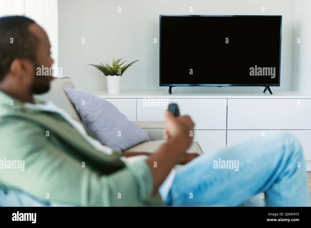 Relaxed black guy watching TV holding remote control and switching channels on plasma television set with blank screen Stock Photo