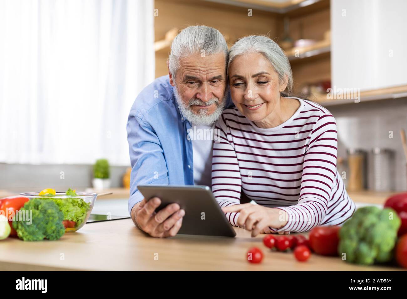 Portrait Of Happy Elderly Spouses Using Digital Tablet In Kitchen Interior, Stock Photo