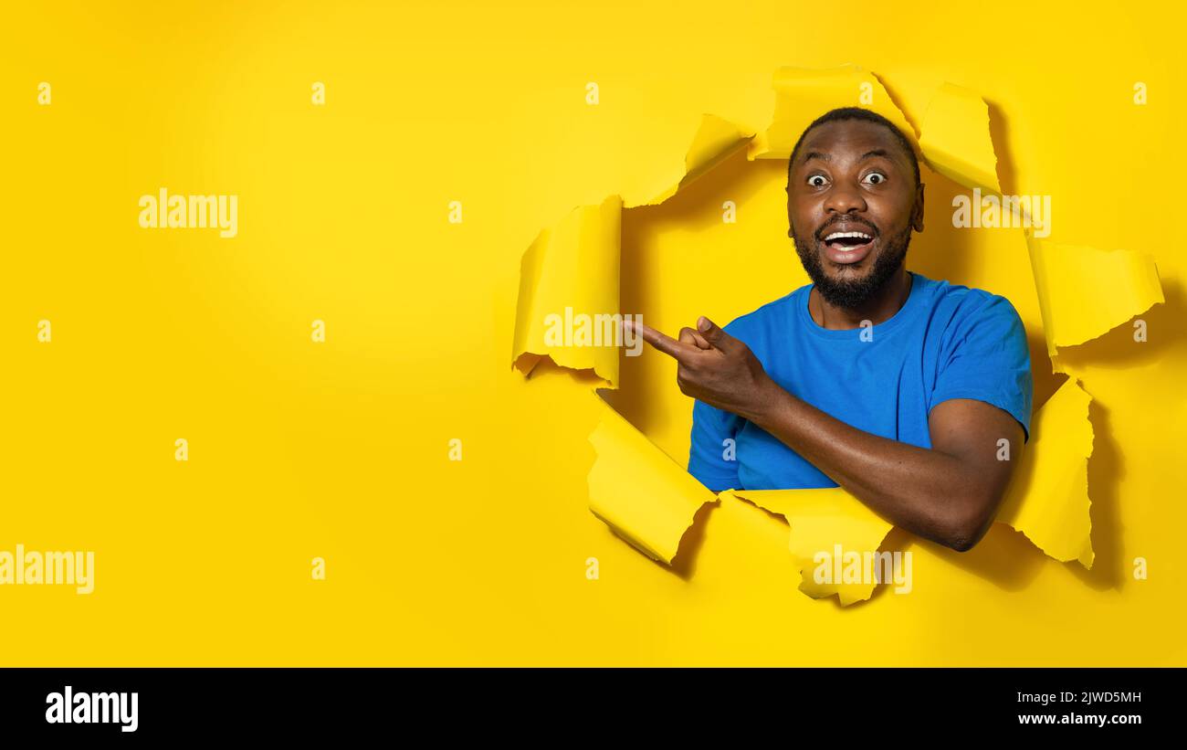 Excited black man gazing at camera with happy expression, pointing at free space, peeking through yellow background Stock Photo