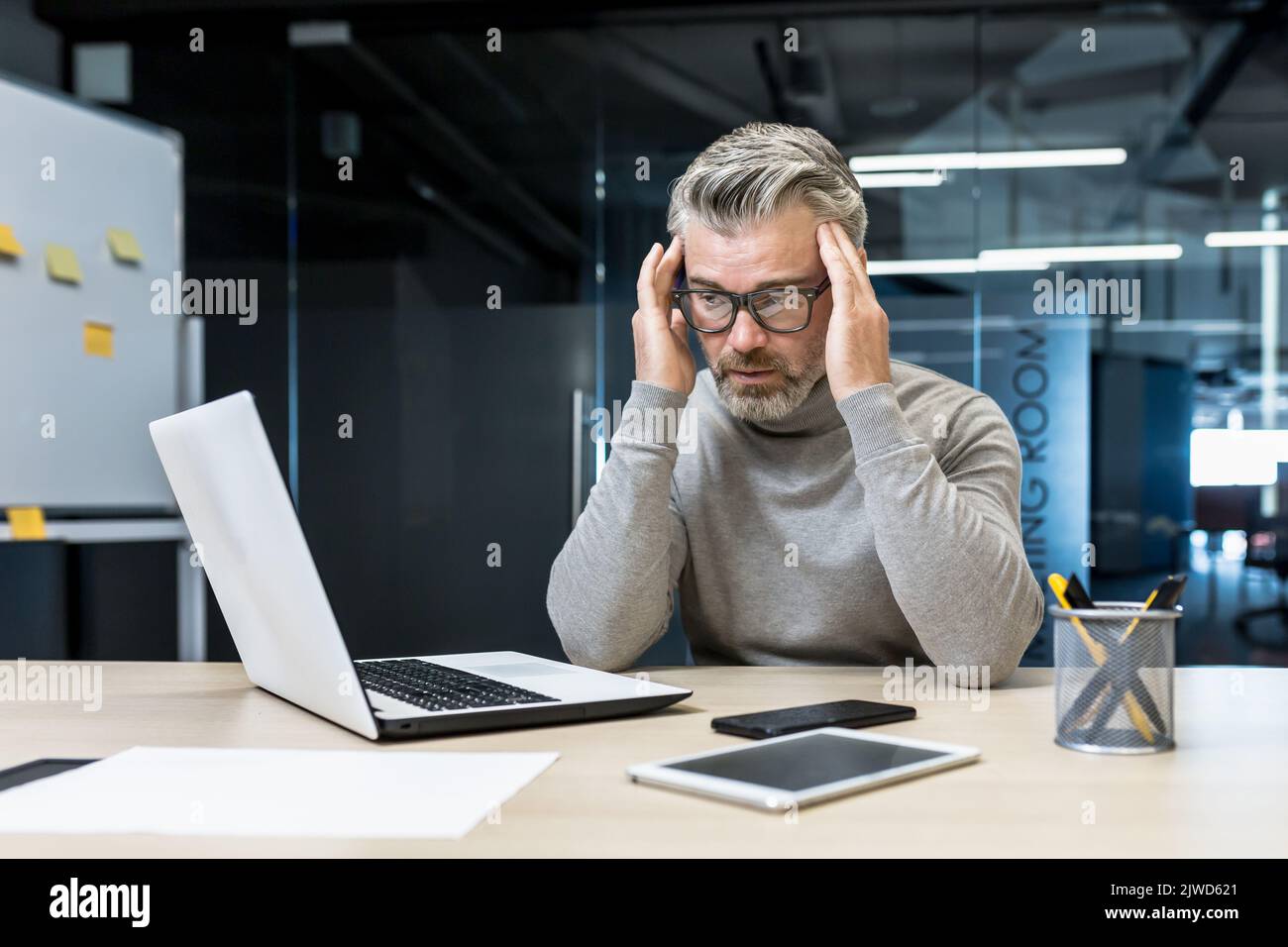 Sick mature businessman working in modern office, senior experienced man having severe headache, manager massaging temples while sitting on chair, manager using laptop for work Stock Photo