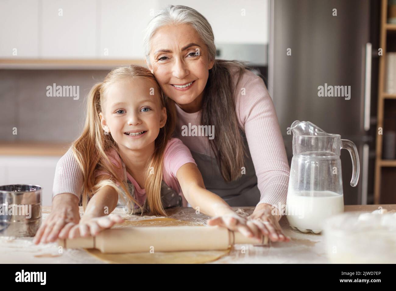 Happy retired woman in apron teaching little girl doing dough using rolling pin making cookies Stock Photo