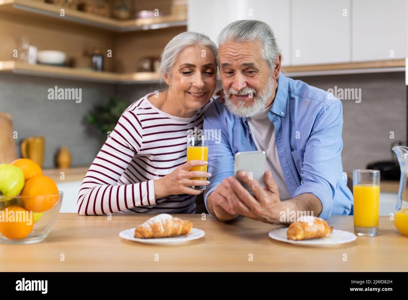 Portrait Of Happy Senior Spouses Using Smartphone During Breakfast In Kitchen Stock Photo