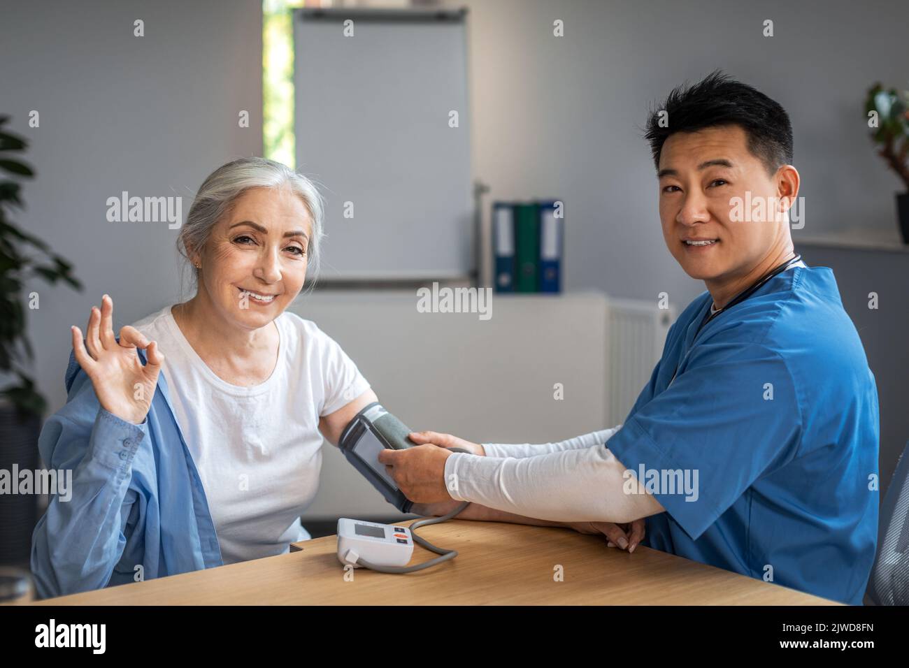 Handsome mature korean man doctor measures pressure with tonometer to senior female, patient waving hand Stock Photo