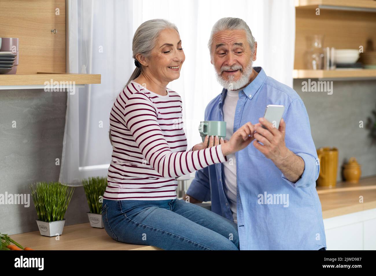 Happy Senior Spouses Using Smartphone Together While Relaxing In Kitchen Stock Photo