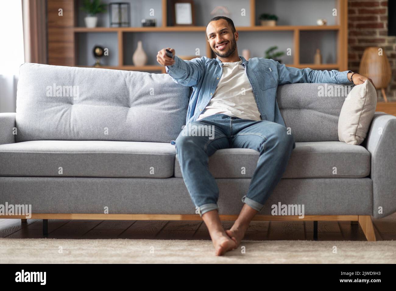 Home Relax. Handsome Black Man Sitting On Couch And Switching Tv Channels Stock Photo