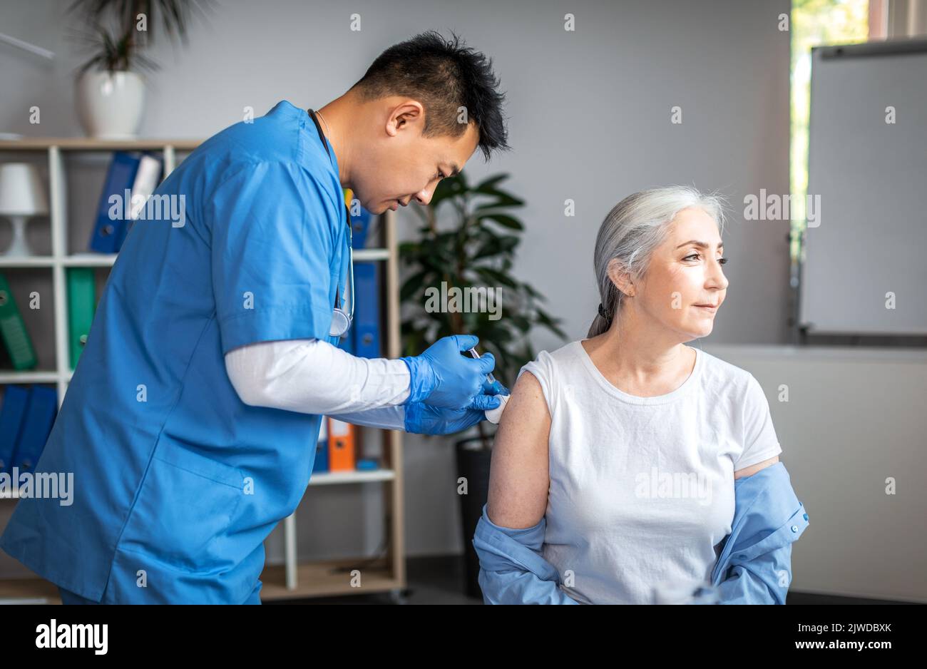 Serious adult asian man doctor do injection to old woman patient in clinic office interior Stock Photo