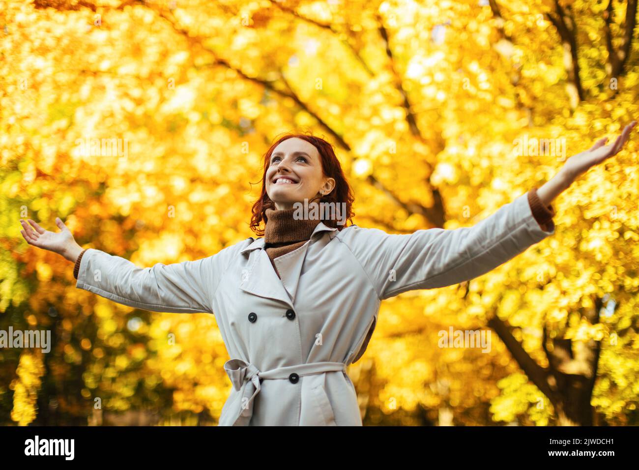 Smiling young european lady in raincoat enjoy good weather, freedom in spare time in park with yellow leaves Stock Photo