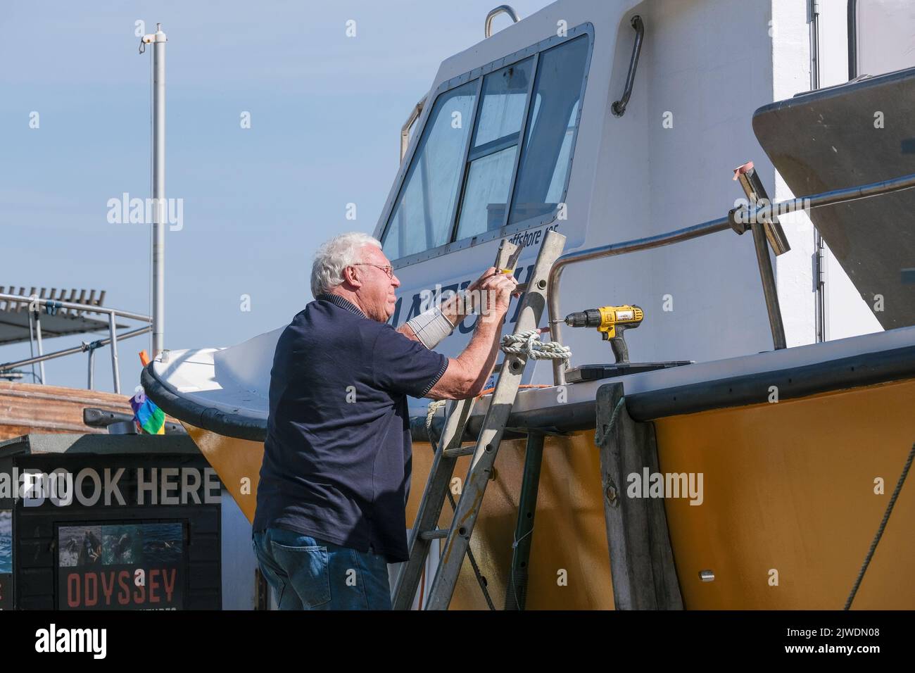 A mature man on a ladder carrying out maintenance on a boat in Newquay Harbour in England in the UK. Stock Photo
