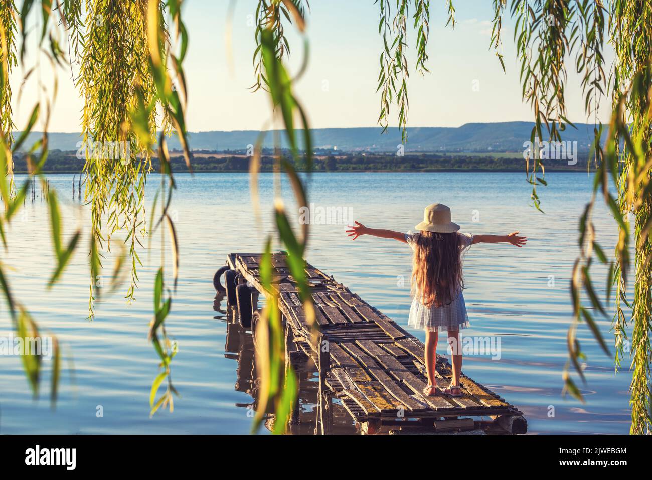 Girl on a old wooden fishing pier and willow tree enjoying beautiful sunset over the sea lake Stock Photo