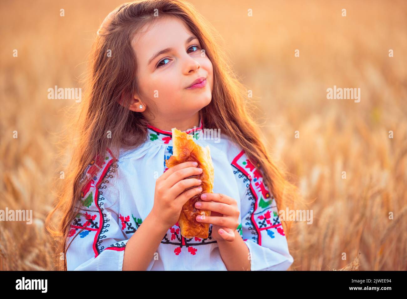 Bulgarian girl, beautiful woman, eating freshly baked banitsa, cheese pie during harvest In golden wheat field Stock Photo