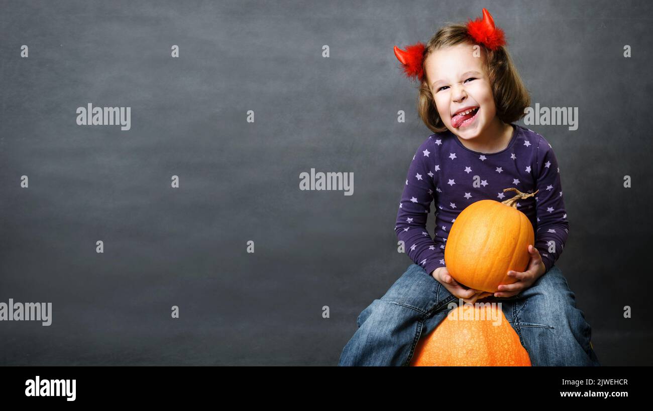 Halloween background with happy kid and space, funny child holds pumpkin and shows tongue, cute little girl with costume horns having fun and makes fa Stock Photo