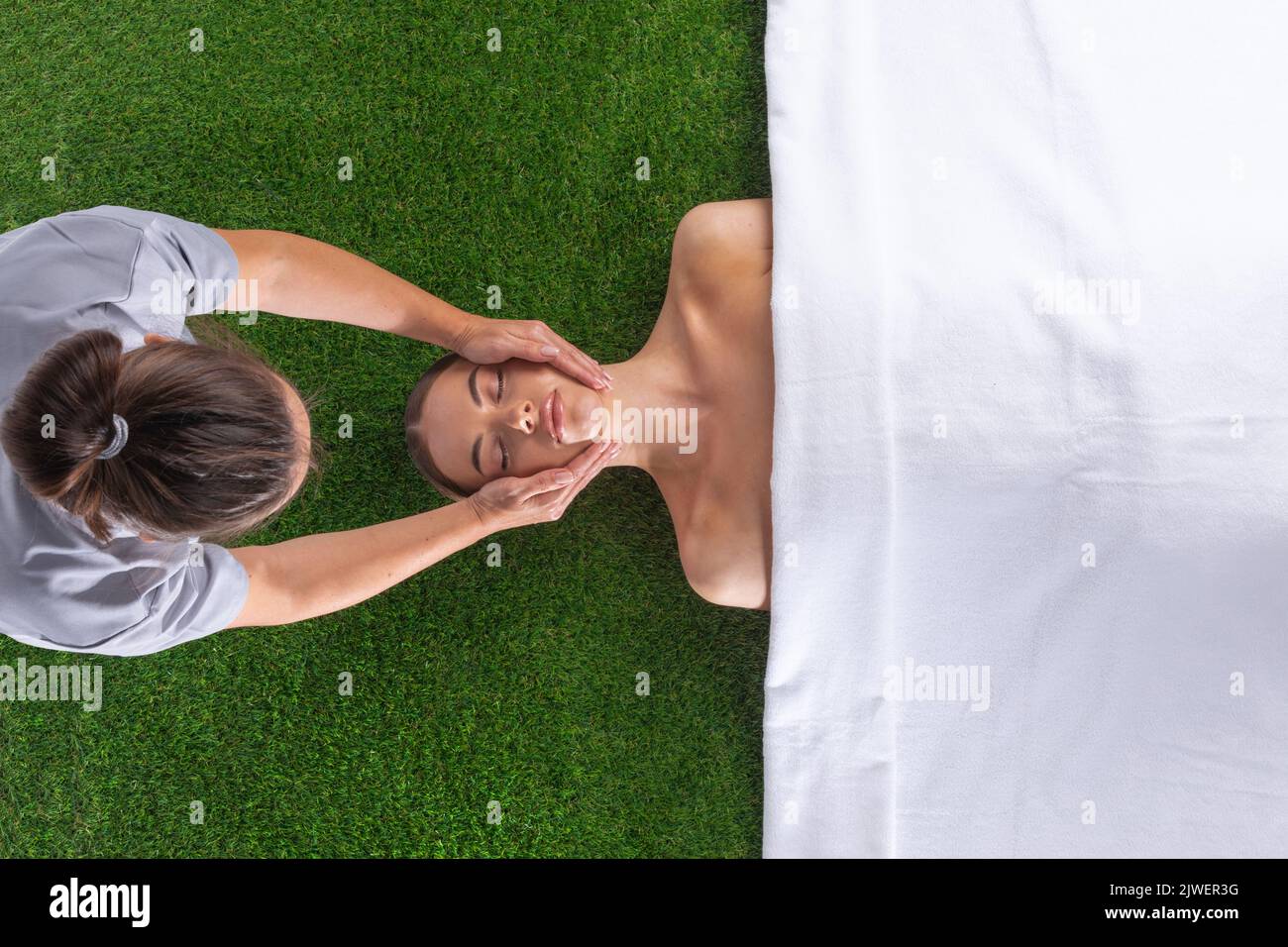 Releasing stress. Top view of beautiful young woman lying on back while massage therapist massaging her face over green grass background Stock Photo