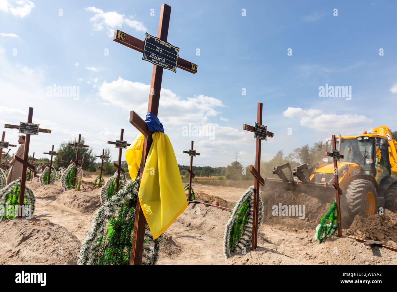 A grave cross tied with a Ukrainian flag seen against the background of an excavator digging pits for fresh graves. Burial of the remains of 13 unidentified and two identified people who were killed in the Buchan district during the Russian occupation, on September 2, 2022. A few months after the de-occupation of the Bucha district, those bodies that were not identified were buried and marked with numbers at the cemetery in Bucha. According to the deputy mayor of Buch Mykhailyna Skoryk, a total of 419 people who were killed during the Russian occupation of the Buchan district were buried in th Stock Photo