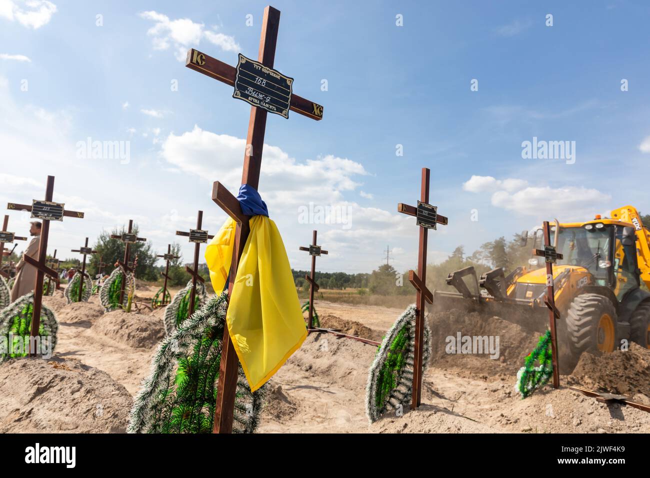 Bucha, Ukraine. 2nd Sep, 2022. A grave cross tied with a Ukrainian flag seen against the background of an excavator digging pits for fresh graves. Burial of the remains of 13 unidentified and two identified people who were killed in the Buchan district during the Russian occupation, on September 2, 2022. A few months after the de-occupation of the Bucha district, those bodies that were not identified were buried and marked with numbers at the cemetery in Bucha. According to the deputy mayor of Buch Mykhailyna Skoryk, a total of 419 people who were killed during the Russian occupation of th Stock Photo