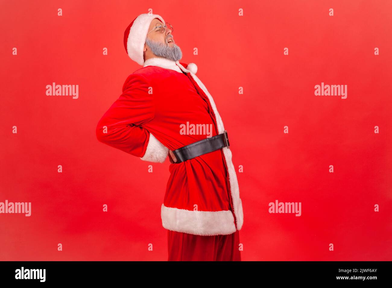 Sick elderly man with gray beard wearing santa claus costume feeling sharp pain in kidneys, back injury, pinched sciatic nerve, suffering. Indoor studio shot isolated on red background. Stock Photo