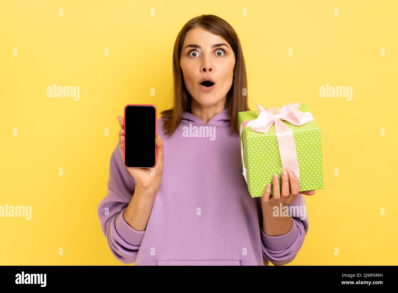 Present, bonus for mobile user. Portrait of amazed surprised woman holding gift box and cell phone with mock up, blank display, wearing hoodie. Indoor studio shot isolated on yellow background. Stock Photo