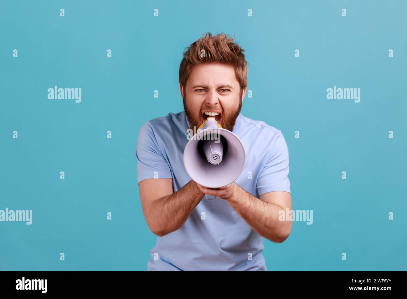Portrait of bearded man holding megaphone near mouth, loudly speaking, screaming, making announcement, paying attention at social problems. Indoor studio shot isolated on blue background. Stock Photo