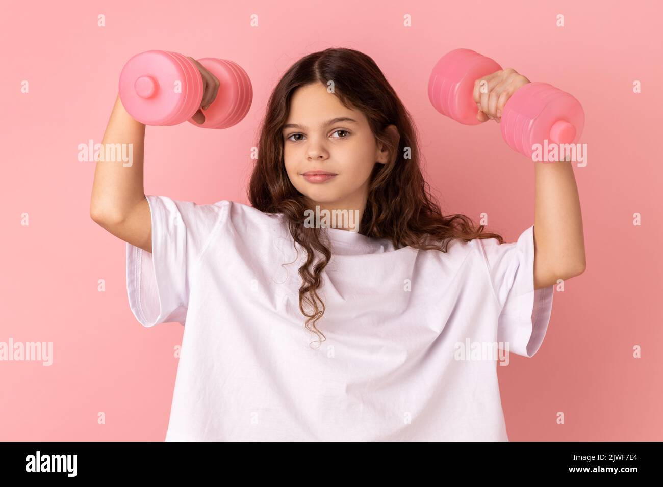 Portrait of strong athletic little girl wearing white T-shirt looking at camera and holding rose dumbbells in raised arms, healthy lifestyle. Indoor studio shot isolated on pink background. Stock Photo