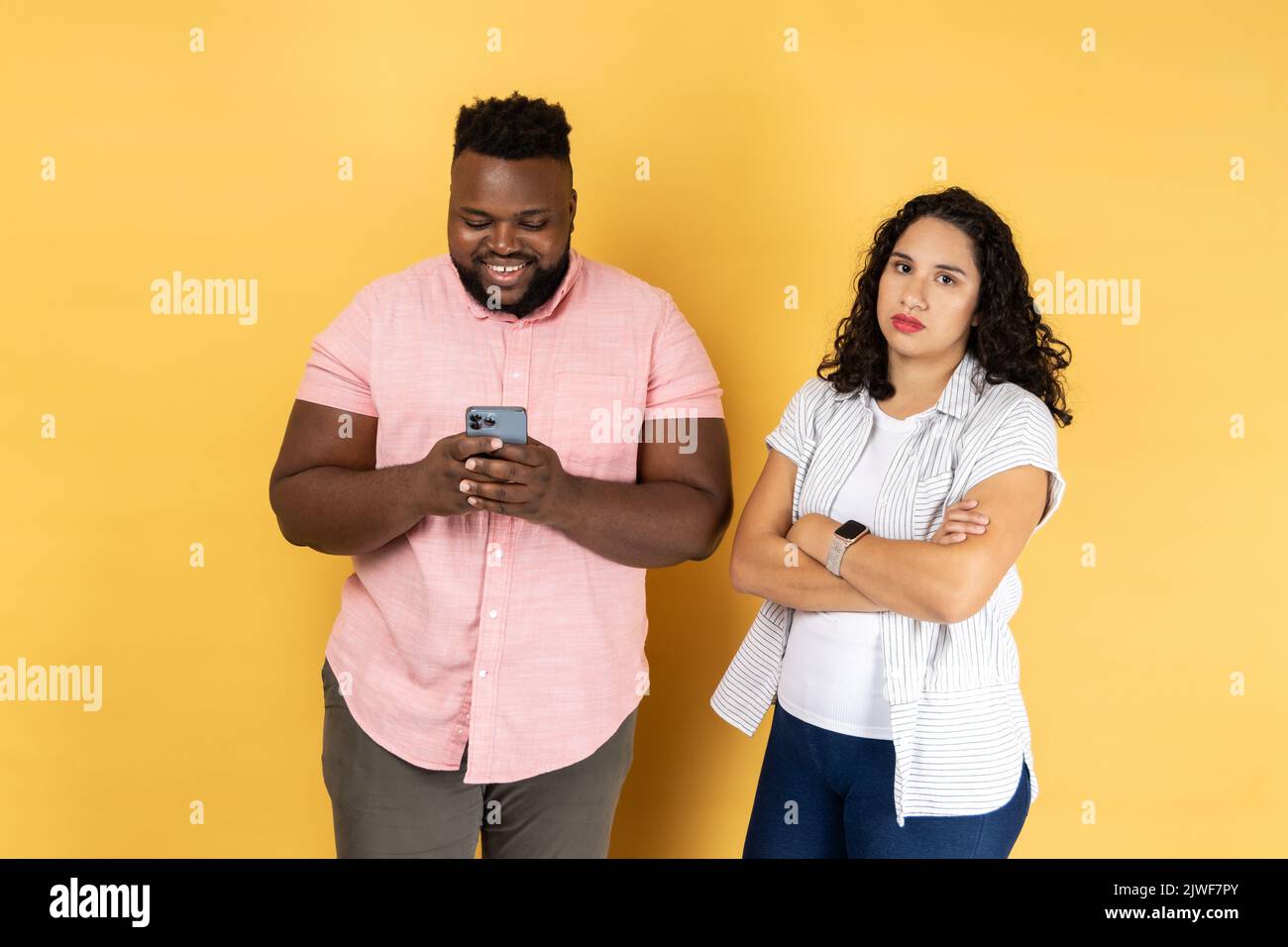 Satisfied young couple in casual clothing standing together, man using mobile phone and smiling, offended lonely woman standing with folded arms. Indoor studio shot isolated on yellow background. Stock Photo