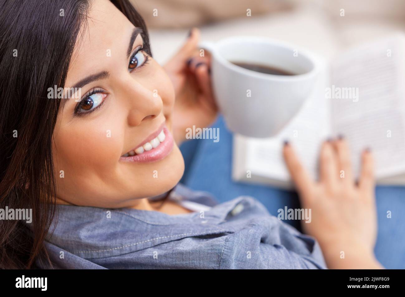 Beautiful happy young Latina Hispanic woman sitting drinking a cup of tea or coffee reading a paperback book at home on her sofa Stock Photo